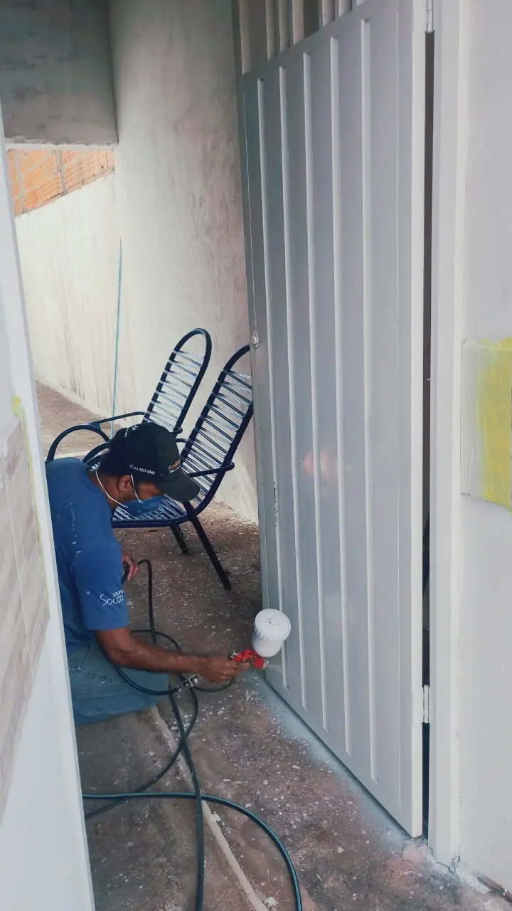 Anderson agachado pintando um portão branco com pistola de pintura, usando boné e máscara.