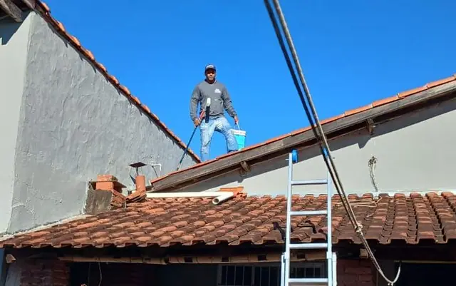 Anderson em pé sobre o telhado, com escada apoiada e balde na mão, lavando a superfície antes da pintura.